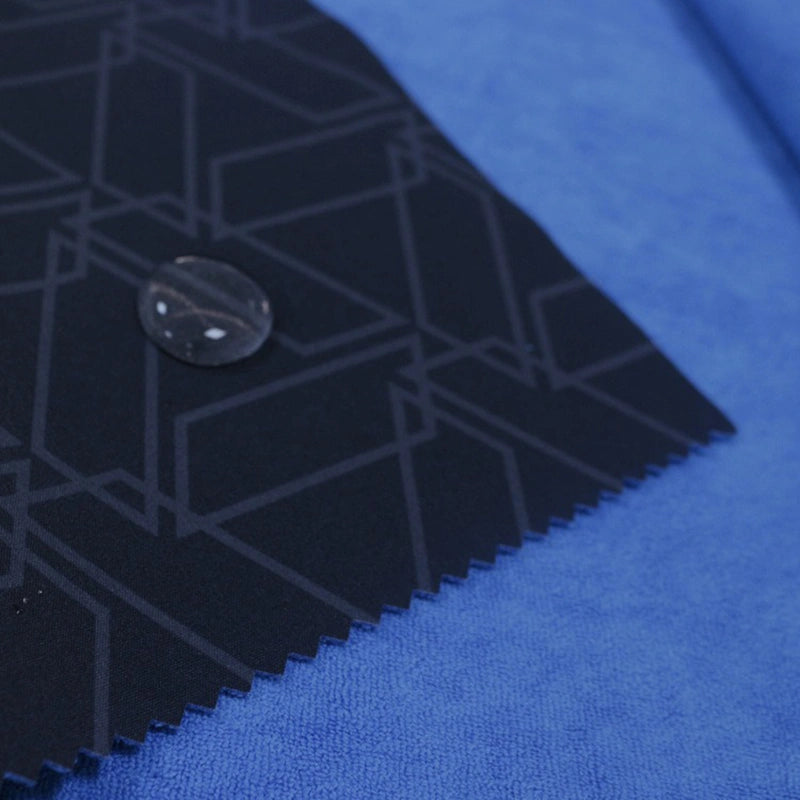 A close-up of two fabric swatches: Longon Craft’s blue Waterproof Polar Fleece Fabric By The Yard beneath a dark, geometric synthetic polar fleece; a black button tops the neat, pinked edge of the upper swatch.