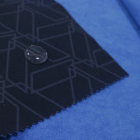 A close-up of two fabric swatches: Longon Craft’s blue Waterproof Polar Fleece Fabric By The Yard beneath a dark, geometric synthetic polar fleece; a black button tops the neat, pinked edge of the upper swatch.