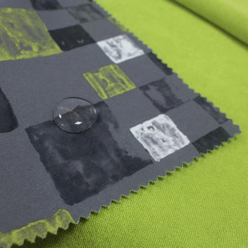 A close-up of Longon Craft’s Waterproof Polar Fleece Fabric By The Yard, featuring a gray pattern with black, white, and yellow squares and a water droplet on the surface, laid over a plain green background.