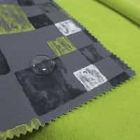 A close-up of Longon Craft’s Waterproof Polar Fleece Fabric By The Yard, featuring a gray pattern with black, white, and yellow squares and a water droplet on the surface, laid over a plain green background.