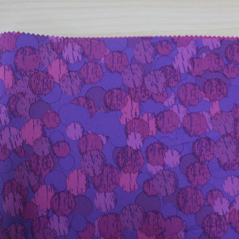A close-up of Longon Craft's Waterproof Polar Fleece Fabric By The Yard shows its purple and pink overlapping circles pattern on a light wooden background, with the polyester fabric's edge cut in a zigzag using pinking shears.