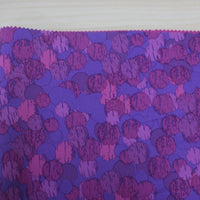A close-up of Longon Craft's Waterproof Polar Fleece Fabric By The Yard shows its purple and pink overlapping circles pattern on a light wooden background, with the polyester fabric's edge cut in a zigzag using pinking shears.