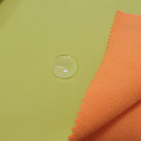 A clear water droplet sits on light yellow Longon Craft Waterproof Polar Fleece Fabric by the yard, beside orange zigzag-edged fabric, illustrating the water resistance and quality of this 4-way stretch synthetic polyester material.